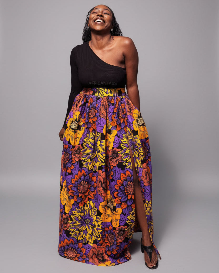 Woman in a colorful skirt and black top standing outdoors with greenery in the background